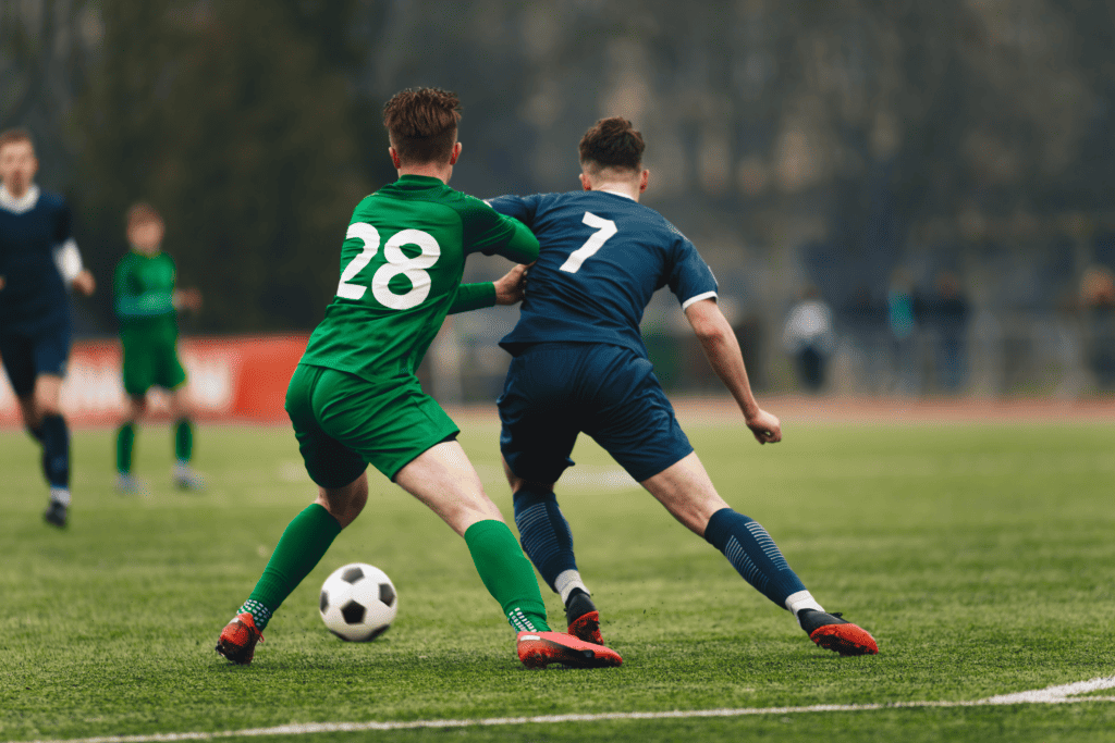 Two college soccer players, one wearing dark blue and the other green, jostling over the ball during a match.