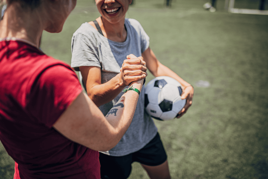 Two women shake hands hands while one holds a black and white soccer ball.