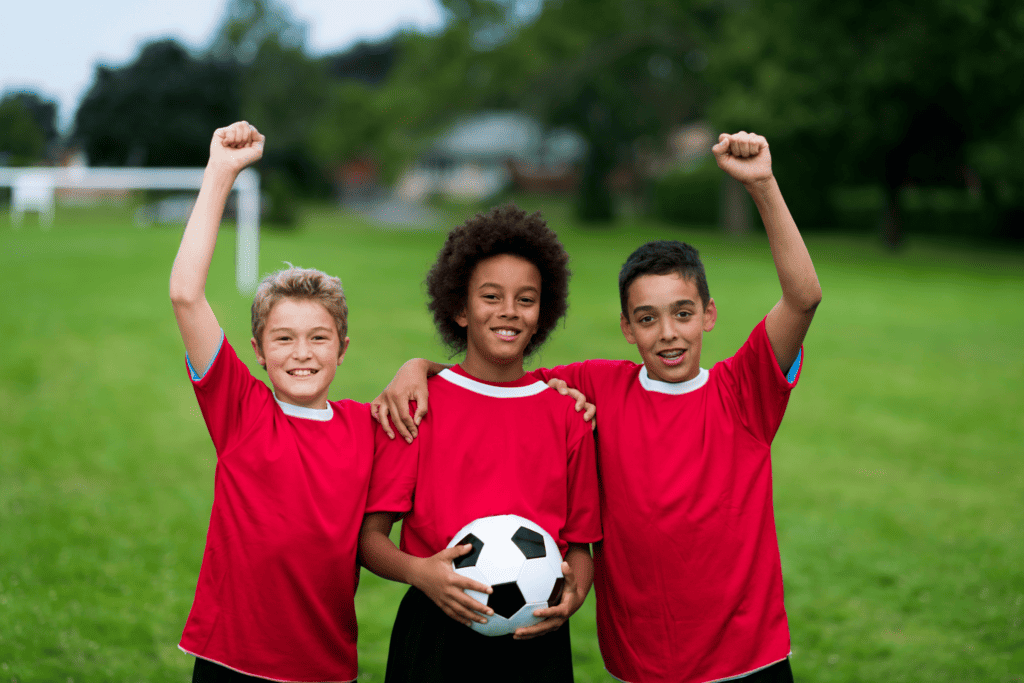 Three youth soccer players stand shoulder-to-shoulder smiling and wearing red shirts. Two raise their fists into the air, while the middle holds a black and white soccer ball.