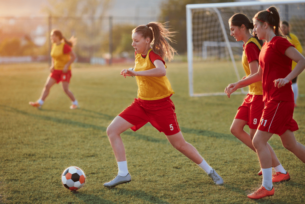 A girl dribbling a ball during a college soccer walk-on tryout wearing red with a yellow athletic bib overtop.